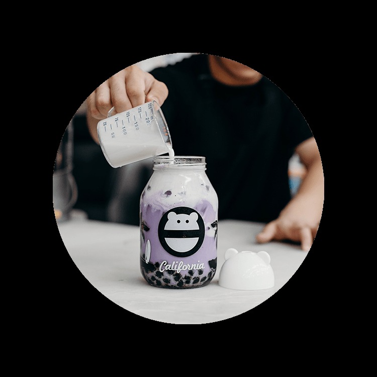 Barista pouring milk from a measuring cup into a taro milk tea in a monster-face glass jar, with a bear-ear lid beside it
