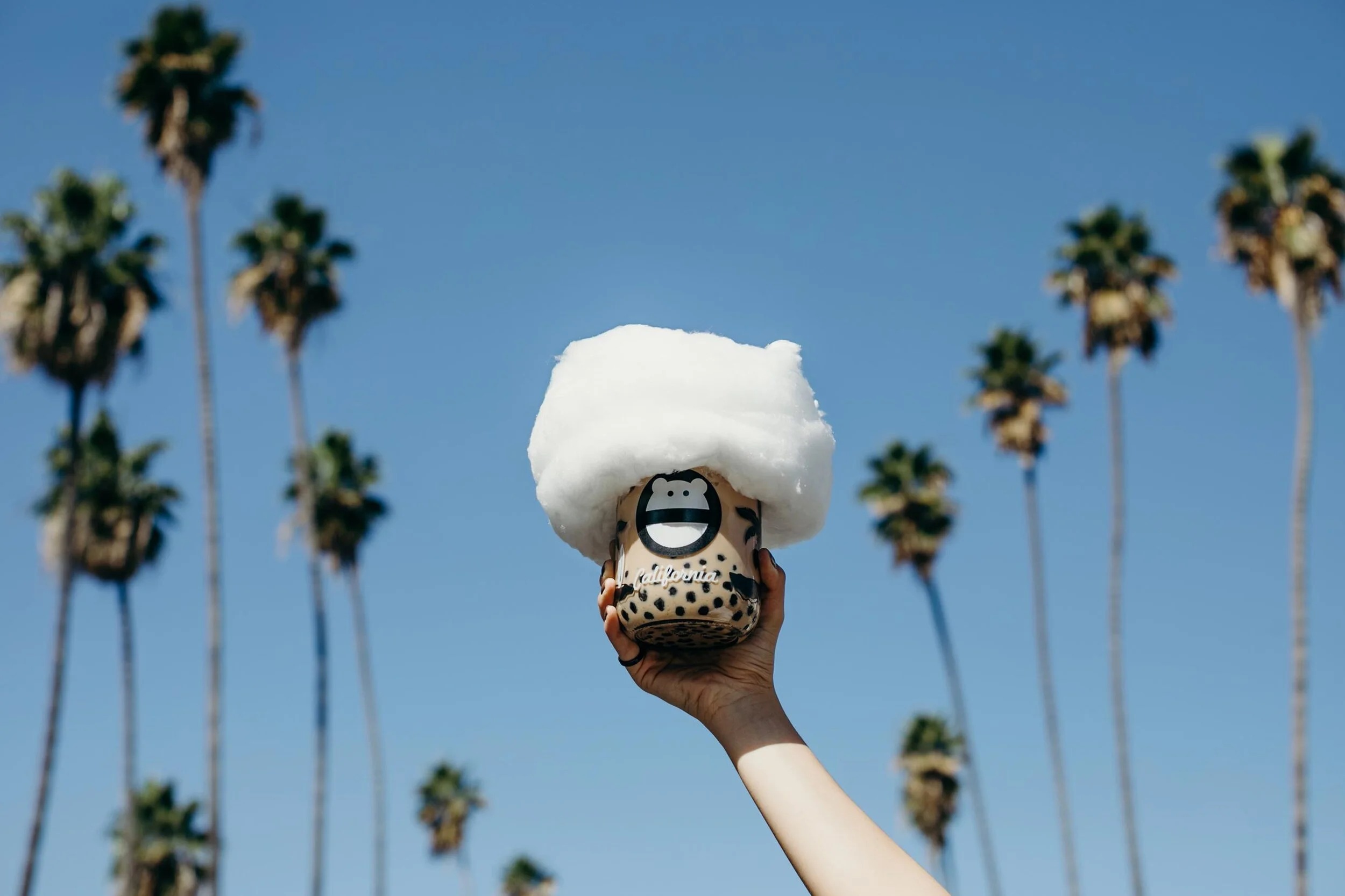 Hand holding a glass “California” jar of jasmine milk tea with boba, topped with a cloud of cotton candy, against a blue sky and tall palm trees