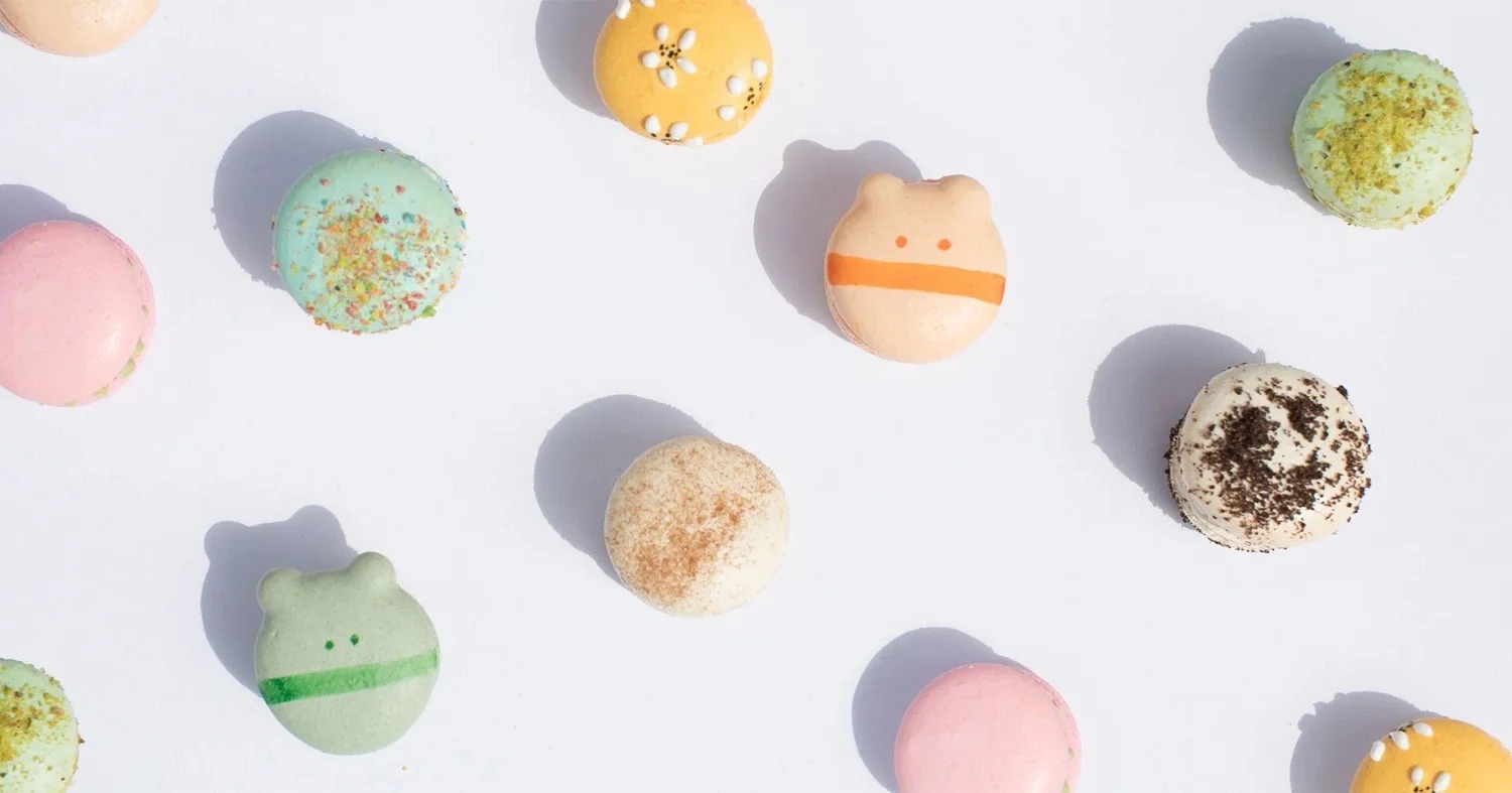 Flat-lay of assorted pastel macarons — pink, mint, daisy-topped yellow, cinnamon, and cookies-and-cream — including bear-eared monster-face macarons in peach and green