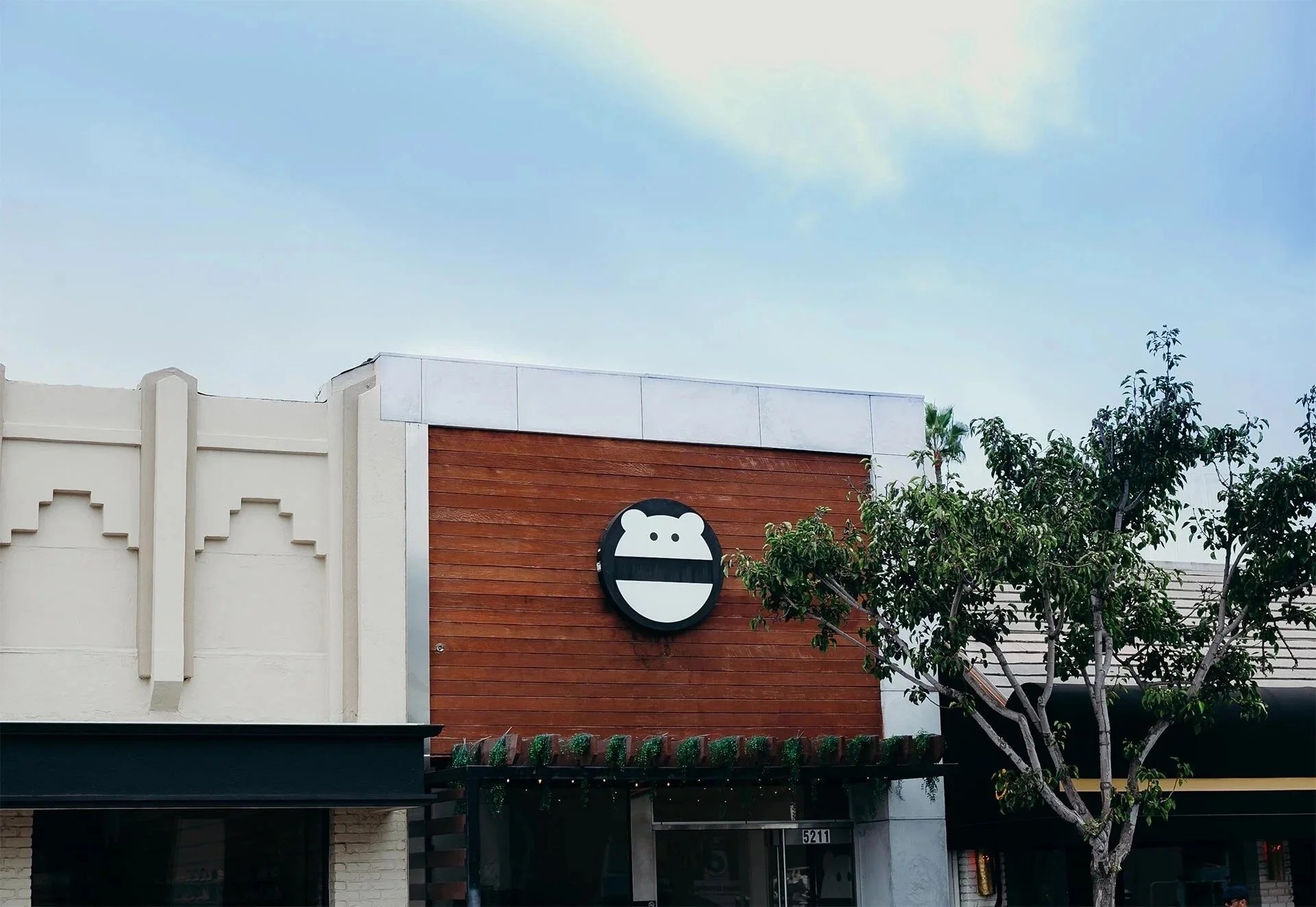 Mini Monster Cafe storefront in Long Beach: wood-paneled facade with a round black monster-face sign, next to an art deco building under a blue sky
