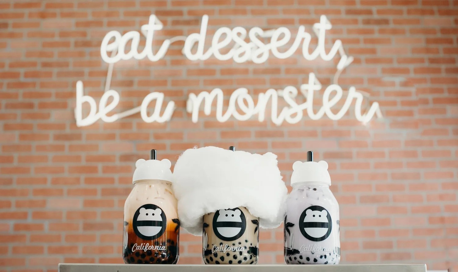 Three boba drinks in monster-face glass jars — thai tea, milk tea topped with cotton candy, and taro — on a counter in front of a brick wall with a white neon sign reading “eat dessert, be a monster”