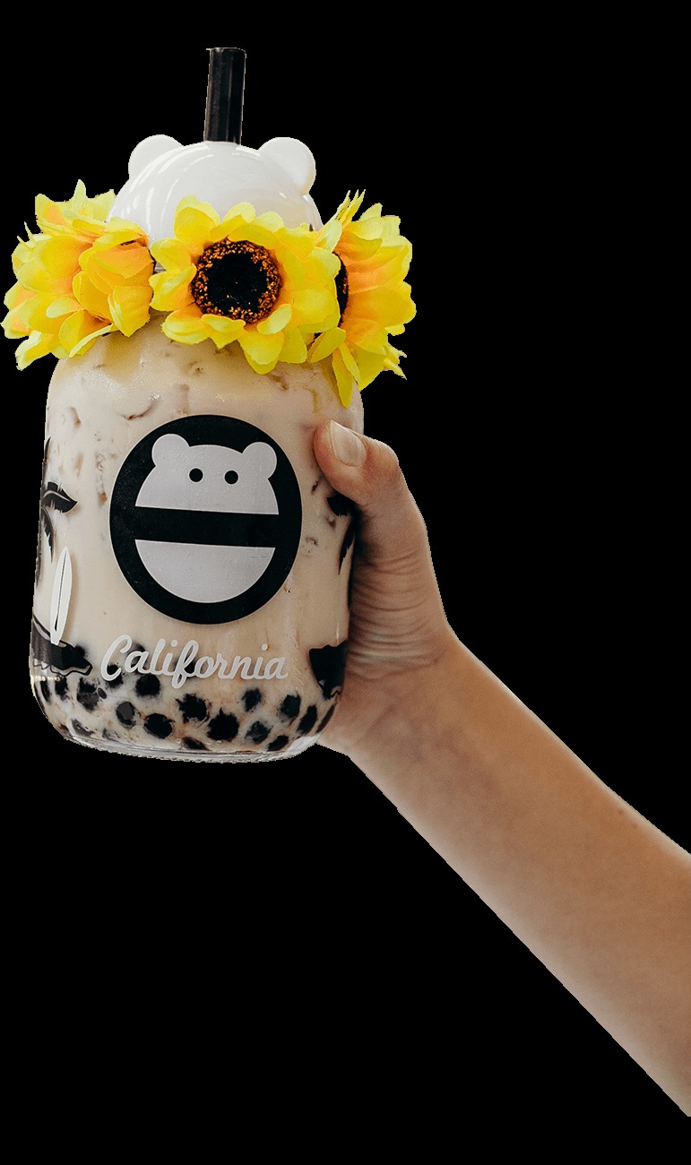 Hand holding a jasmine milk tea with boba in a monster-face “California” glass jar with a bear-ear lid and black straw, wreathed in yellow sunflowers