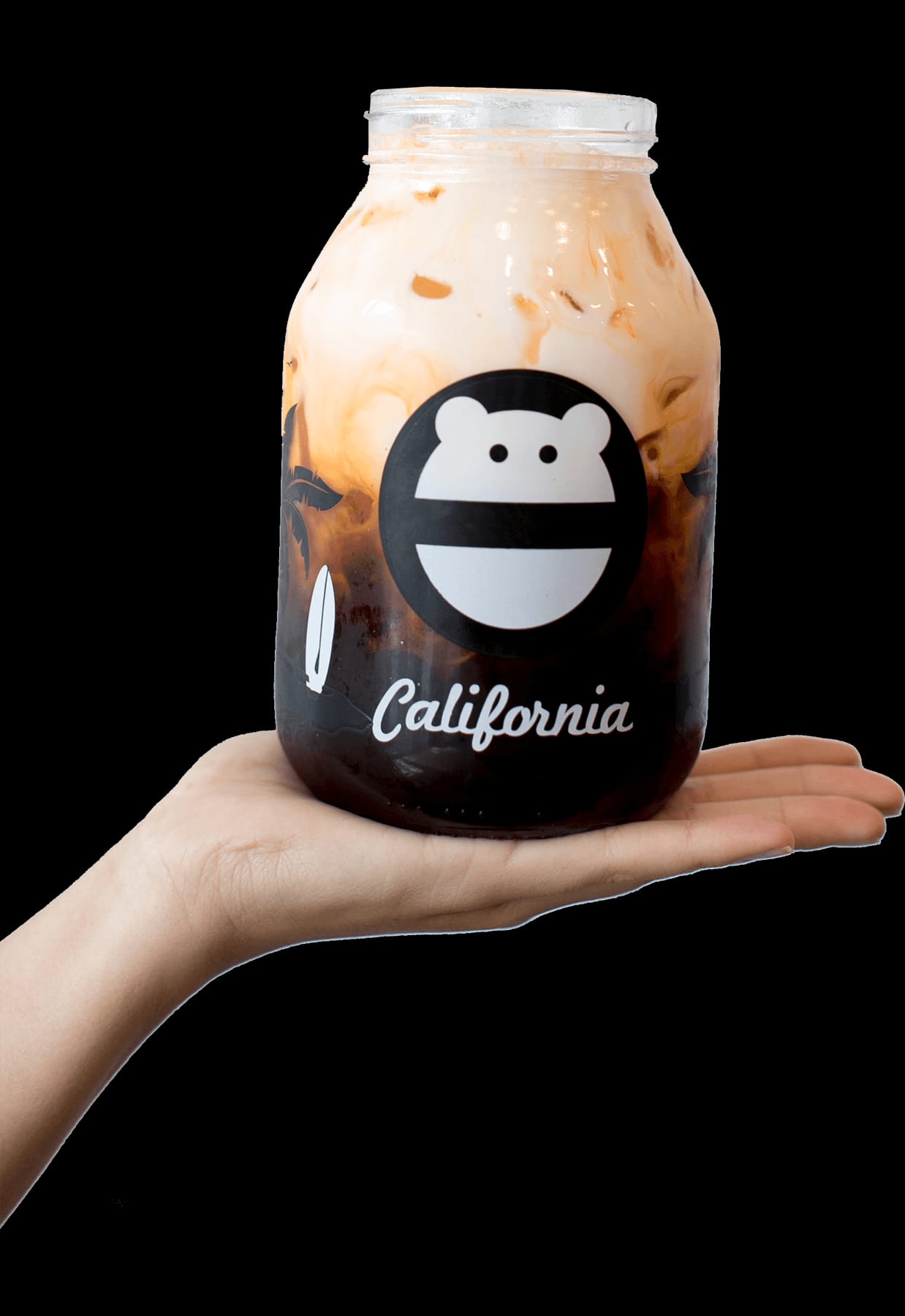 Open palm holding a reusable glass “California” jar of thai tea with cream swirling into the dark tea, etched with the monster face logo and surfboard and palm graphics